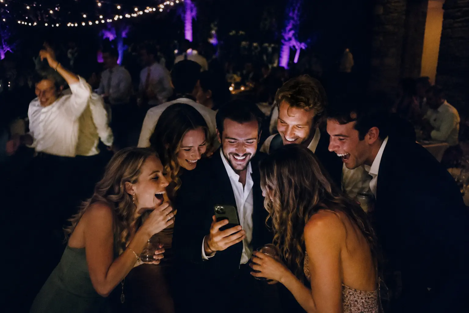 A huddle of wedding guests laughing around one glowing phone on a dark dance floor.
