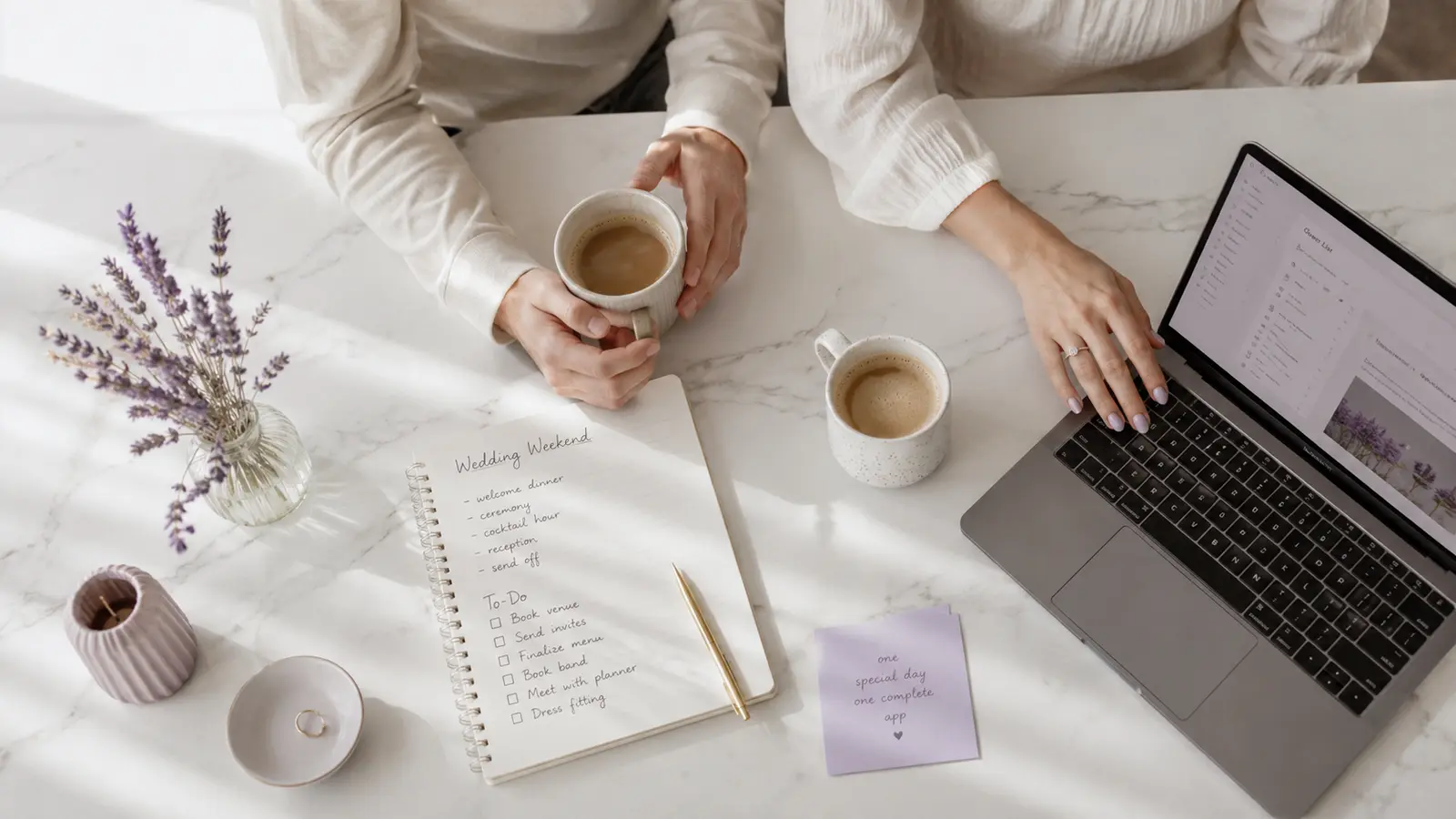 A couple at a bright kitchen table with coffee, a wedding notebook, and their guest list open on a laptop.